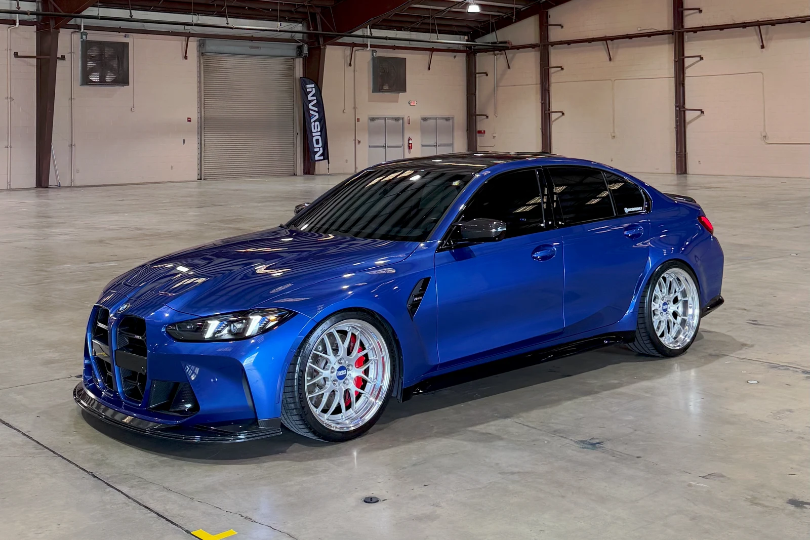 Blue BMW G80 M3 on white forged wheels with carbon front lip inside a warehouse