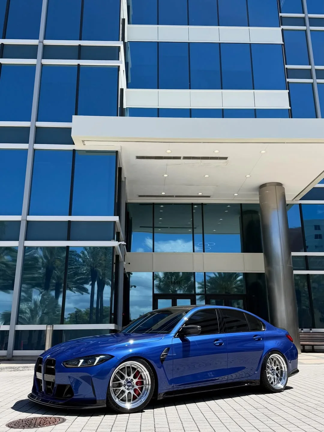 Blue G80 M3 on polished wheels beneath a glass tower